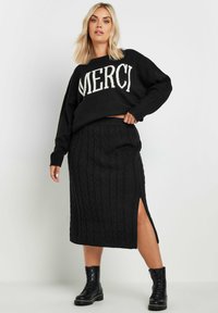 Mustat neuleetakki, jossa on valkoiset "MERCI"-teksti, yhdistetty mustiin kabelineulehameeseen, jossa on sivurako ja mustat maiharit.