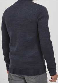 Homme portant un pull en laine tricoté bleu foncé et un jean gris, vu de dos sur un fond gris clair uni.