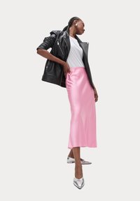 Giacca da moto in pelle nera, t-shirt bianca aderente, gonna midi in raso rosa e ballerine argentate con punta. Modella in posa di profilo.