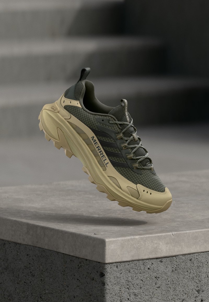 Αθλητικό παπούτσι για τρέξιμο σε εξωτερικούς χώρους με μπεζ σόλα και σκούρο πράσινο πλέγμα στο επάνω μέρος, αιωρείται πάνω από μια άκρη από σκυρόδεμα, με τη σήμανση "Merrell" στο πλάι.