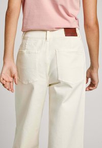 Pantaloni beige a vita alta con una vestibilità rilassata. Presentano due tasche posteriori e un'etichetta in pelle marrone sulla vita.