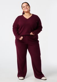 Lachende vrouw in een bordeauxrood geribbeld V-hals trui en bijpassende broek, met de zoom van de trui in haar hand, en witte gestikte schoenen.