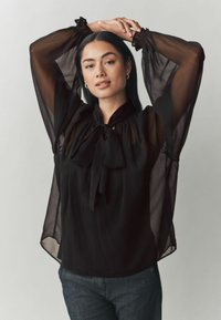 Čierna priesvitná blúzka s dlhými, vzdušnými rukávmi sťahovanými pri zápästiach. Obsahuje viazaný golier a hladkú textúru, skombinovaná s tmavými džínsami.