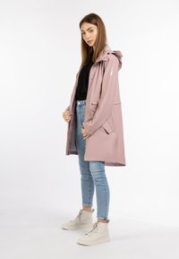 Lys pink vandtæt regnfrakke med hætte, sølvdetaljer, snøre i taljen og frontlommer, båret over en sort top og blå jeans.