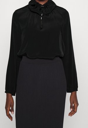 Blouse noire à manches longues avec col montant froncé et cordon, associée à une jupe côtelée foncée, portée par une personne des épaules aux hanches.