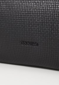 Sac à main en cuir synthétique noir texturé avec un motif en grille subtil et le logo "Calvin Klein" embossé à l'avant.