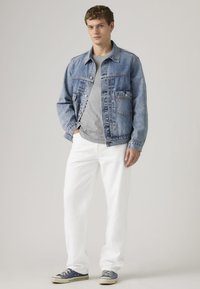 Νέος άνδρας που στέκεται με τα χέρια στις τσέπες, φορώντας μπλε τζιν μπουφάν, γκρι μπλούζα, λευκό παντελόνι και σκούρα μπλε αθλητικά παπούτσια σε φωτεινό φόντο.