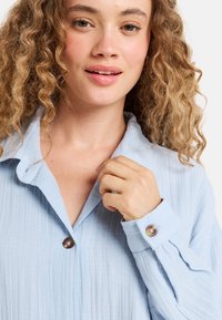 Lichtblauw, getextureerd overhemd met een gekraagde halslijn, knoopdetails en een relaxte pasvorm. De stof valt zacht en heeft ribbelpatronen.