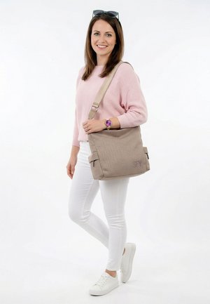 Femme en pull rose et pantalon blanc souriante, portant un sac à épaule beige, des baskets blanches et des lunettes de soleil sur la tête.