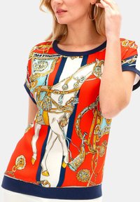 Czerwono-niebieska bluzka w wzory przedstawiające motywy jeździeckie, krótkie rękawy i prążkowany dekolt. Gładki materiał z złotymi akcentami i detalami.