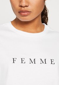 Witte katoenen T-shirt met dikke zwarte blokletters die het woord "FEMME" spellen. Ronde halslijn en korte mouwen, minimalistisch ontwerp.
