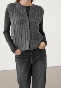 Graue Strickweste mit Zopfmuster, rundem Ausschnitt, fünf Knöpfen und geripptem Saum, getragen über einem Langarmshirt und kombiniert mit grauen Jeans.