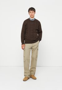 Maglione marrone a treccia, pantaloni cargo beige con tasche e sneaker color sabbia. L'outfit presenta texture a contrasto e una vestibilità rilassata.