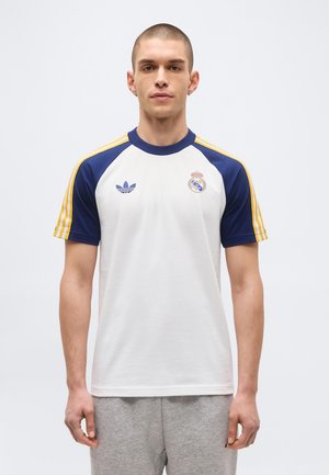 Jonge man draagt een wit Adidas-Real Madrid T-shirt met marineblauwe mouwen en gele strepen, staand tegen een effen lichte achtergrond.