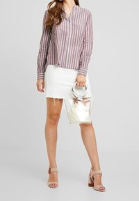 Chemise à rayures mauve et blanche, boutonnée, associée à une jupe en denim blanc et un sac transparent. Chaussures à talons avec bride à la cheville.