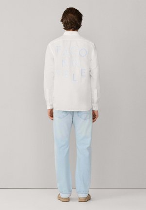 Hombre con camisa blanca de manga larga con el texto "FACONNABLE" en la espalda, jeans azul claro y zapatos beige sin cordones, de espaldas.