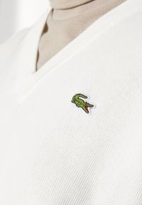 Witte v-hals trui van textiel met een groene krokodillenlogo geborduurd op de linkerkant van de borst.