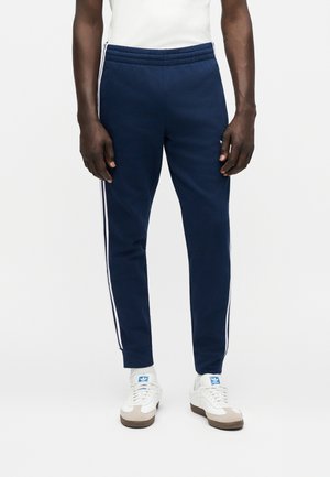 Mężczyzna noszący granatowe dresowe spodnie z białymi paskami po bokach, białe skarpety i białe buty sportowe Adidas z niebieskim logo.