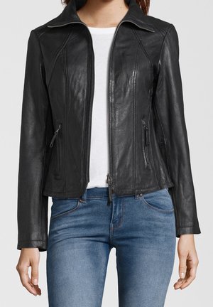 Vrouw draagt een zwarte leren jas met rits over een wit shirt en blauwe jeans, staand met ontspannen handen langs haar zij.