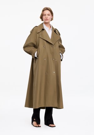 Femme portant un long manteau croisé vert olive sur une chemise blanche et un pantalon noir fendu à l'ourlet, avec des sandales noires à bout ouvert.