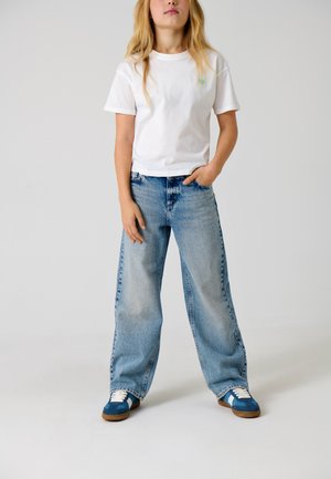 Jeune personne aux longs cheveux blonds portant un t-shirt blanc, un jean bleu clair ample et des baskets bleues, debout avec une main dans la poche.