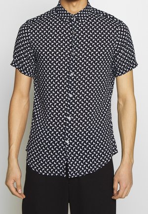 Mann trägt ein schwarzes Kurzarmhemd mit weißem geometrischem Muster und schwarze Hose vor einfachem Hintergrund.