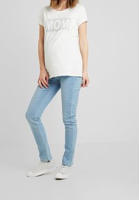 Těhotenské grafické tričko v krémové barvě s designem "MOM", spárované se světle modrými skinny džínami a šedými teniskami s bílou podrážkou.