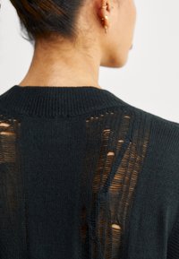 Svart stickad tröja med ribbad krage, med distinkta horisontella och vertikala utskärningar längs ryggen för en extra textur.