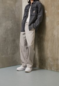 Šedá džínová košile s dvěma kapsami na hrudi, bílý tílko, béžové kalhoty a bílé tenisky. Pozadí tvoří betonová zeď.