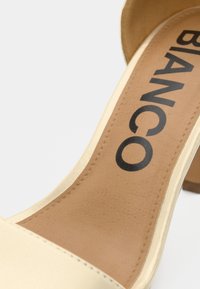 Μπεζ πέδιλο με τακούνι, με απαλό υφασμάτινο επάνω μέρος, καφέ υποδήμα από τεχνητό δέρμα, και έντονη γραφή "BIANCO". Διαθέτει τακούνι τύπου μπλοκ.