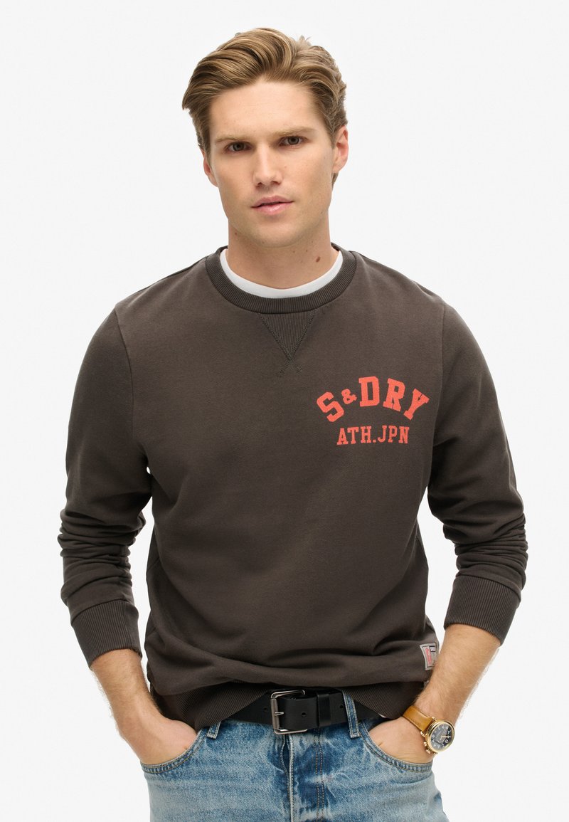 Sudadera marrón de cuello redondo con texto rojo "S&D DRY ATH.JPN", puños y dobladillo acanalados. Llevada con jeans de mezclilla azules y un cinturón negro.