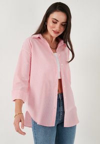 Pink and white striped button-up shirt with a collar, rolled sleeves, and side slits, paired with a white top and blue jeans.