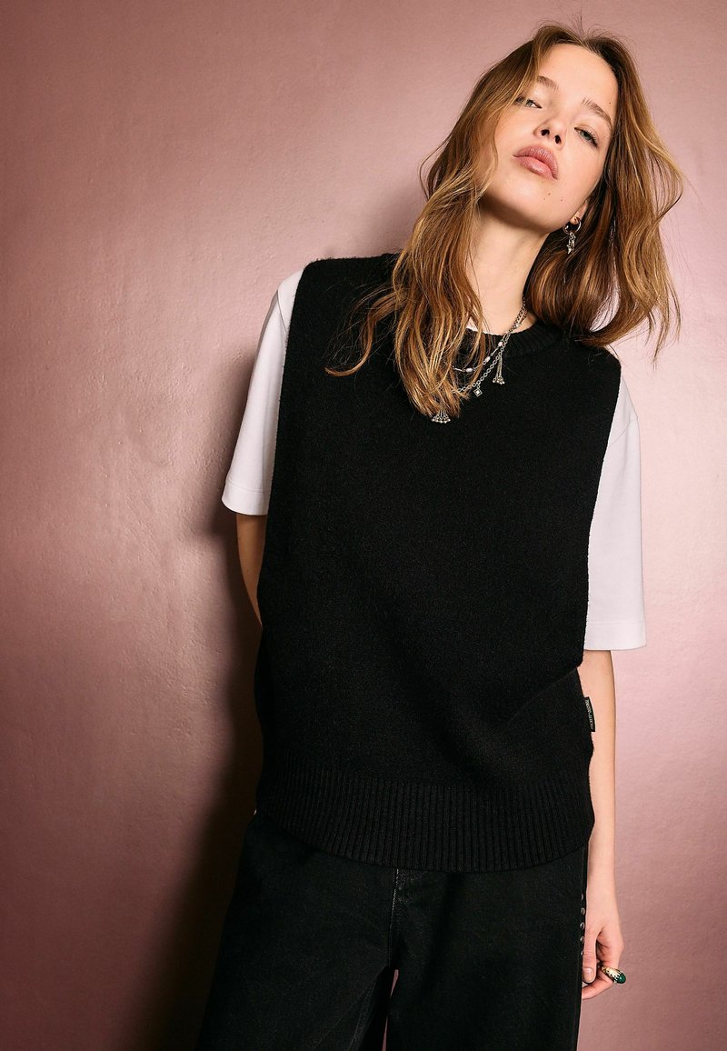 Fiatal nő világosbarna hajjal, fekete ujjatlan pulóvert visel fehér rövid ujjú ing fölött, egy visszafogott rózsaszín fal előtt áll.