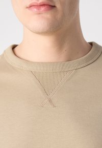 Bežová mikina s ribovaným výstrihom a stehmi na golieri. Mäkká textúra, tesne priliehajúci dizajn, minimalistická.