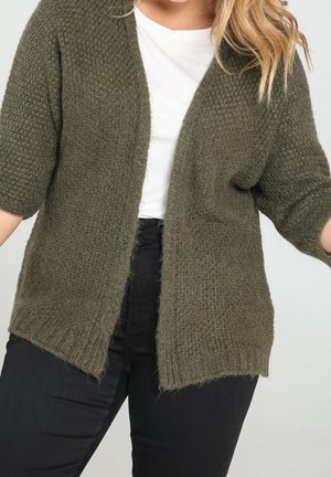 Femme portant un cardigan en maille vert olive ample sur un haut blanc et un pantalon noir, debout devant un fond uni.
