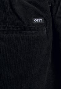 Black corduroy pants with prominent vertical stripes, featuring a small white label stitched on the back pocket displaying "OBEY."