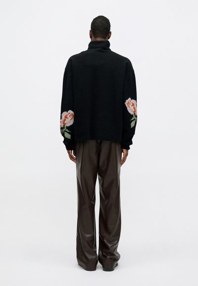 Bărbat stând cu spatele, îmbrăcat cu un pulover negru cu guler înalt, având modele florale pe mâneci și pantaloni din piele maro închis, largi.