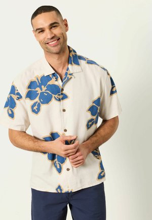 Lächelnder Mann steht in beige kurzärmligem Hemd mit großen blauen Blumenmustern und dunkelblauer Hose vor einem schlichten hellen Hintergrund.