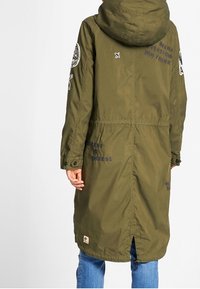 Olivgrön parka med huva, prydd med broderad text och patchar, tryckknappar och en åtdragen midja. Halvlång med en ryggslits.