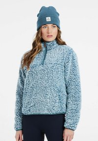 Pullover in pile azzurro chiaro con colletto alto e zip frontale a metà. Presenta una superficie texture e fluffy e polsi a costine. Abbinato a un berretto coordinato.