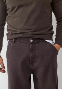 Bruine trui en donkerpaarse broek. De broek heeft een knoopsluiting en zijzakken. De materialen lijken zacht met een gladde textuur.