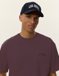 Maroon oversized t-shirt with navy embroidered logo on the chest, paired with a navy cap featuring white text "LES DEUX." Simple, minimal design.