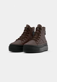 Brązowe skórzane wysokie sneakersy z grubą czarną podeszwą, zaokrąglonym noskiem i sznurowanym wzorem. Posiadają naszywkę z logo z boku.