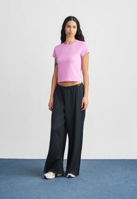 Różowy T-shirt z krótkim rękawem z logo, zestawiony z czarnymi plisowanymi sportowymi spodniami. Model ma na sobie białe buty sportowe. Prosty i funkcjonalny design.