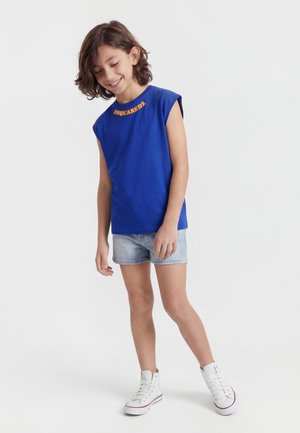 Smilende barn med skulderlangt brunt hår iført en blå ærmeløs top, lyse denimshorts og hvide høje sneakers.