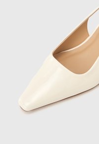 Escarpin à bout pointu en cuir verni crème brillant avec une texture lisse et un dos ouvert, présentant un design minimaliste.