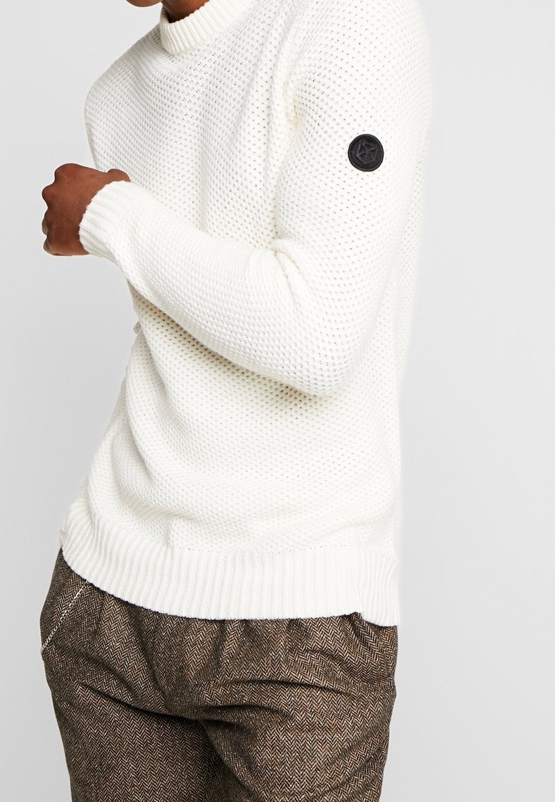 Homme portant un pull en maille blanche texturée avec un patch circulaire noir sur la manche et un pantalon marron à motifs, le bras plié sur la poitrine.