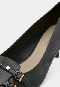 Salgado preto de pele com biqueira pontuda, apresentando uma alça com detalhes em metal prateado e dourado. Textura suave e design de salto baixo.