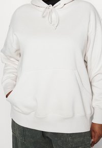 Světle béžová mikina vyrobená z měkké tkaniny, s klokaní kapsou, kapucí na šňůrku a ribovanými manžetami a lemem.