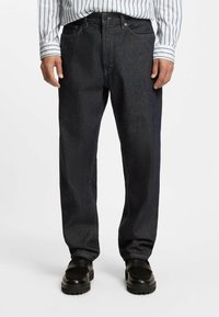 Jeans in denim blu scuro con taglio a gamba dritta, caratterizzati da tasche frontali e dettagli di cucitura sottili. Indossati con mocassini neri e una camicia a righe.